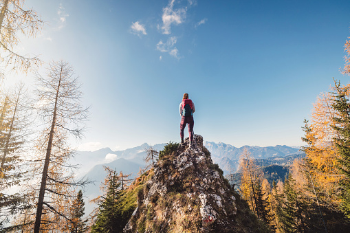 Scenic view of autumn mountain, European Alps, from a view point, where caucasian woman hiker is standing. Sun is shining high up in the mountains, a light mist in the valleys down bellow. Woman mountaineer enjoying the view of majestic Alps on a sunny autumn day.