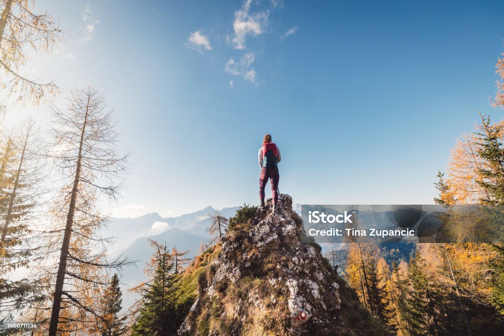 On top of the world - Caucasian woman hiker standing on a rock, admiring the view of autumn mountains in the distance Scenic view of autumn mountain, European Alps, from a view point, where caucasian woman hiker is standing. Sun is shining high up in the mountains, a light mist in the valleys down bellow. Woman mountaineer enjoying the view of majestic Alps on a sunny autumn day. High Tatras Stock Photo