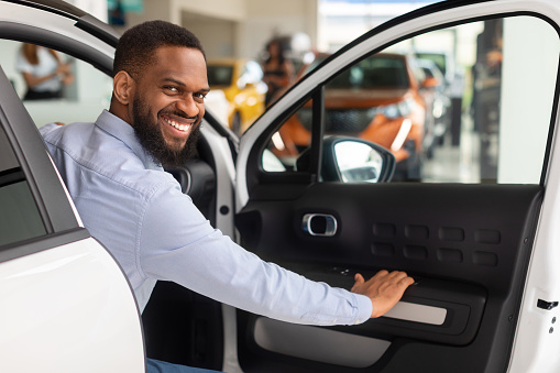 Smiling African American Man Opening Door Of His New Car And Smiling At Camera, Happy Young Black Male Posing In Luxury Vehicle After Purchasing Automobile In Modern Dealership Center, Closeup