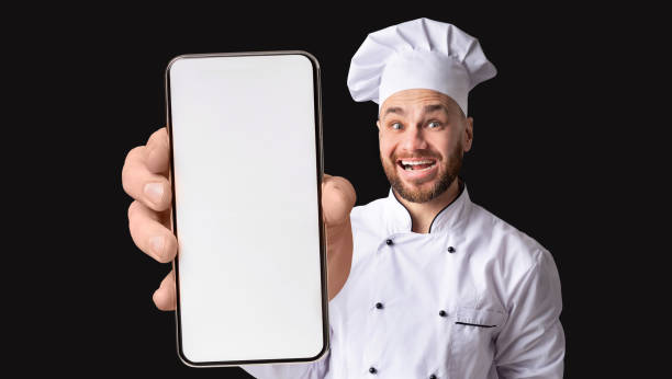 istock Funny Young Chef Holding Demonstrating Smartphone With Blank Screen At Camera 1450713776