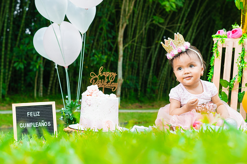 baby brunette latina, sitting on the lawn looking happy, celebrating her first birthday, next to a cake with a happy birthday sign.
