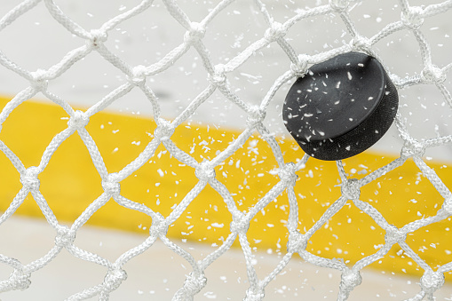 A close-up view of an Ice Hockey puck hitting the back of the goal net as shavings fly by in mid-air. Scoring a goal in ice hockey.