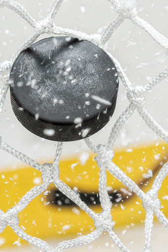 A close-up view of an Ice Hockey puck hitting the back of the goal net as shavings fly by in mid-air, with a hockey stick in the background. Scoring a goal in ice hockey.