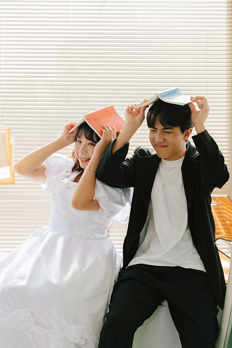 Young Asian heterosexual couple smiling and holding books on top of their head and making cute funny faces while reading. The couple is spending time together in living room relaxing, sitting in front of the window with blinds or curtains under warm sunlight. The woman is  wearing a long white dress.\n\nYoung Asian man male groom and woman female bride in romantic relationship taking pre-wedding photos in Taiwan. A waist up portrait photo about concept of love and romance.\n\nYoung Asian boyfriend and girlfriend enjoying the day and spending time together at home, getting excited about future life plans.