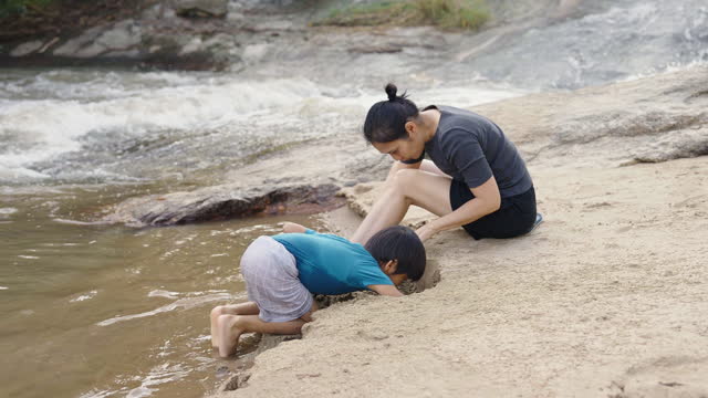 Mother and son digging in the sand on the riverbank near waterfall