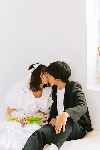 Young Asian heterosexual couple kissing each other lightly, while sitting next to each other on the bed against the white wall under warm sunlight. The woman is holding some flowers in her hand and wearing a white dress and a flower headband.\n\nYoung Asian man male groom and woman female bride in romantic relationship taking pre-wedding photos in Taiwan. A photo about concept of love and romance.\n\nYoung Asian boyfriend and girlfriend enjoying the day and spending time together at home, getting excited about future life plans.