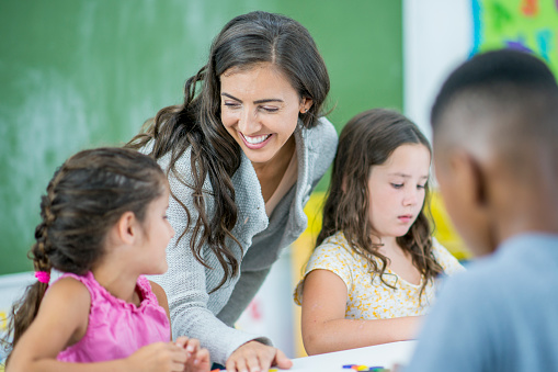A young female teacher of Hispanic decent, leans in to help a student with her work.  She is dressed semi-casually and has a smile on her face as she encourages the student and makes her way around the classroom,