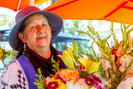 Mature female Ecuadorian Chola Cuencana Florist's portrait with colorful flowers looking at camera at Flower's Market in Cuenca, Ecuador, Latin America / South America. She's wearing a typical Cuenca's handcraft andes clothes.