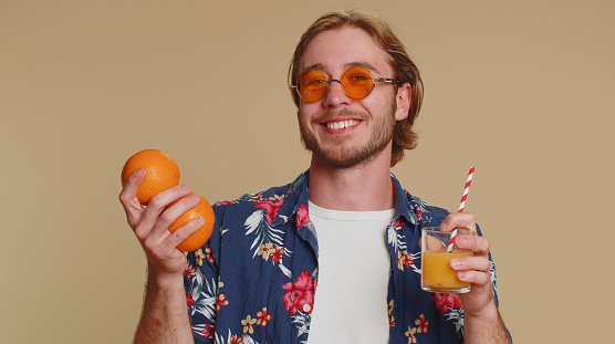 Tourist young man holding, showing oranges fruits, vegetarian lifestyle, vitamins for health, happy vacation holidays, organic eco food, drinking juice from straw. Young guy boy on beige background