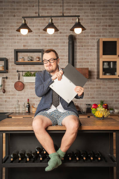 Angry handsome man wearing suit and underpants working at home online siting on the kitchen table and smashing his laptop stock photo