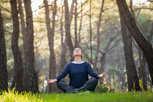 After fitness workout a young woman doing meditation sitting in natural area surrounded by trees, fresh green grass and mountains. She is wearing fitness dress. Her clothes are wet with sweat.