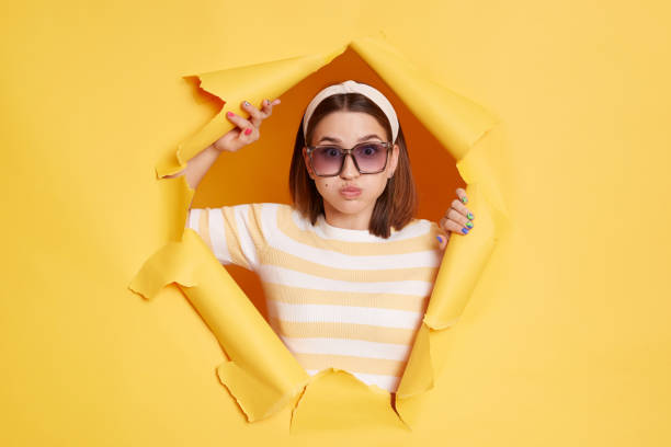 retrato de una divertida mujer tonta con camiseta a rayas, banda para el cabello y gafas de sol, rompiendo el agujero de papel en la pared amarilla, respirando, soplando las mejillas, mirando a la cámara. - chubby cheeks fotografías e imágenes de stock