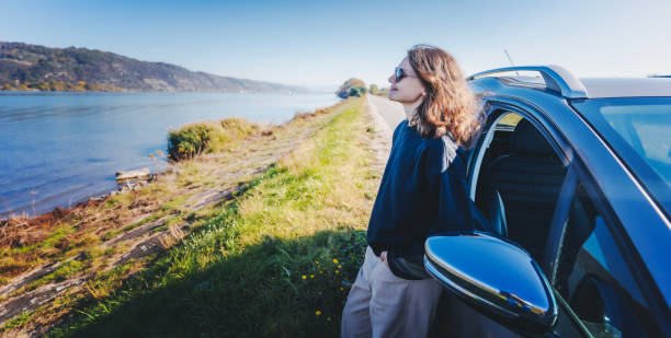 Young cheerful woman traveler standing next to car enjoying sunny day stock photo