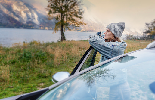 Young cheerful woman driver in a hat and jacket traveling by car standing next to the car in the autumn forest