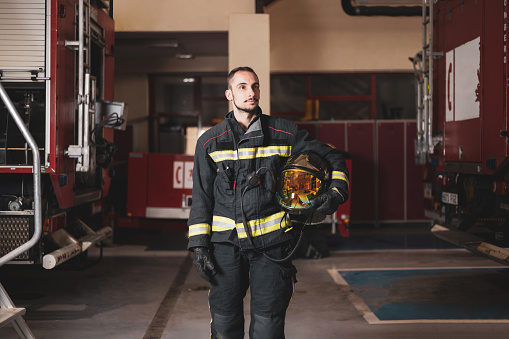 A young firefighter posing in uniform and helmet in hand inside the fire station.