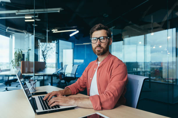 istock Portrait of a successful programmer inside the office, a man with a beard, glasses and a red shirt looks at the camera, a developer engineer uses a laptop to write code in a programming framework 1450770286