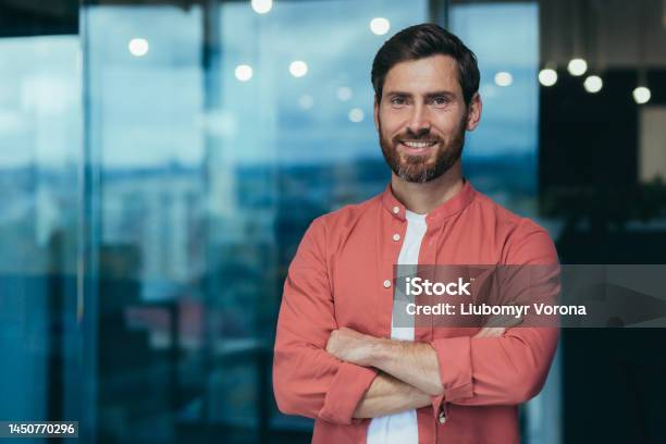 Portrait Of A Happy And Smiling Programmer In The Middle Of A Modern Office A Man In Glasses And A Red Shirt Is Looking At The Camera With His Arms Crossed Stock Photo - Download Image Now