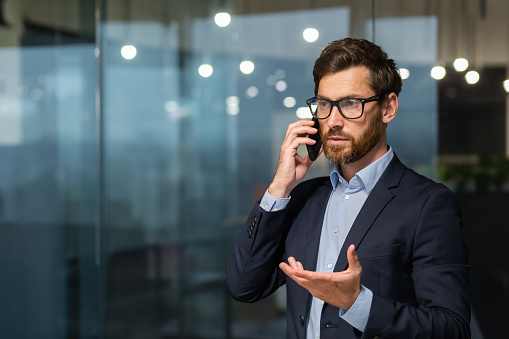 Serious and successful mature businessman inside office near window talking on phone, man in business suit and beard standing.