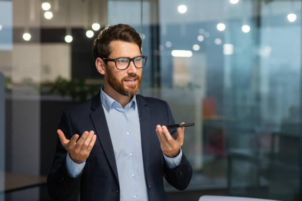 istock Friendly and smiling businessman in office recording audio message, man using smartphone app for communication, investor in business suit near window inside building 1450770360