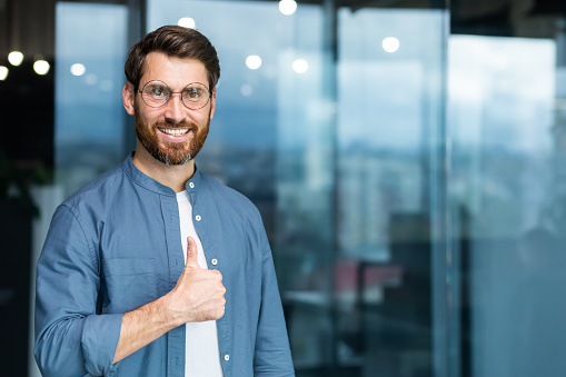 Portrait of businessman in casual shirt, man with beard and glasses smiling and looking at camera showing thumbs up affirmative, business owner working inside office.