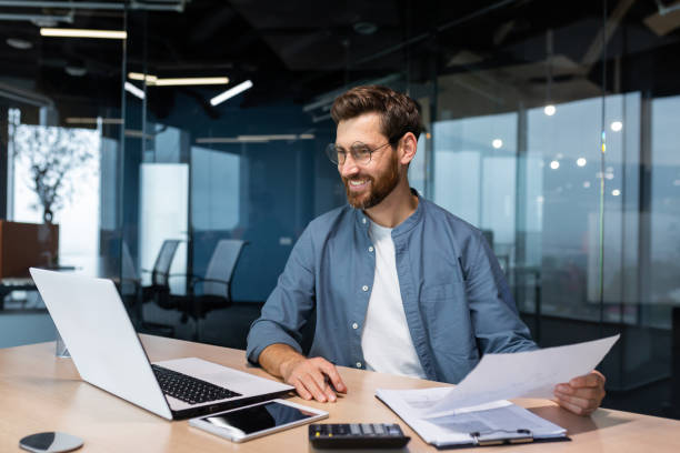 istock Mature businessman in shirt doing paperwork, man working with documents, contracts and bills sitting at table using laptop at work, financier accountant with beard and glasses 1450770594