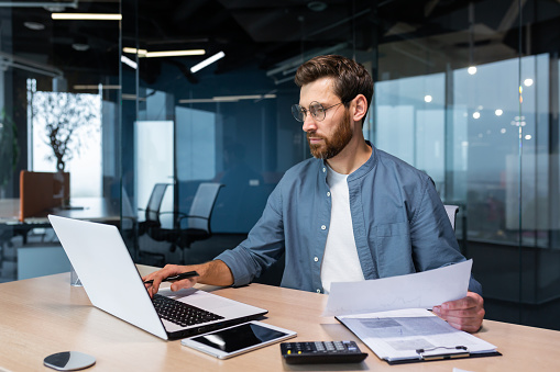 Serious and focused financier accountant on paper work inside office, mature man using calculator and laptop for calculating reports and summarizing accounts, businessman at work in casual clothes.