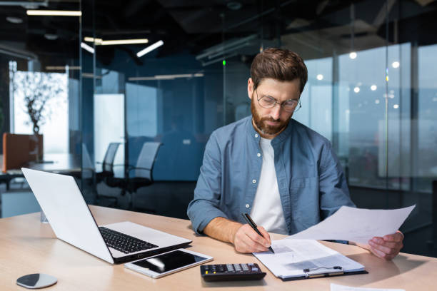 istock Serious and focused financier accountant on paper work inside office, mature man using calculator and laptop for calculating reports and summarizing accounts, businessman at work in casual clothes 1450770697