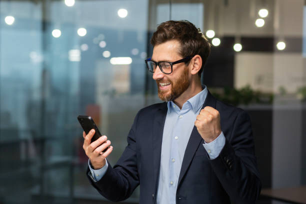 istock Successful businessman investor using phone, man celebrating victory and successful achievement, holding hand up triumph gesture, boss in business suit inside office standing near window 1450770778