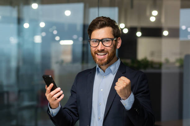 portrait d’un investisseur homme d’affaires prospère, patron mature en costume et lunettes regardant la caméra et heureux de célébrer le succès de la victoire, l’homme tenant un smartphone a reçu en ligne des nouvelles de bonne réalisation - working smiling equipment car photos et images de collection