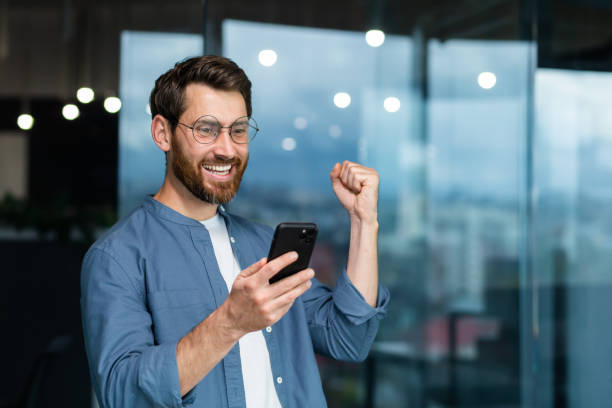 istock Businessman in shirt near window using phone , mature man with beard received notification online good news, worker celebrating victory rejoices, holding hand up gesture of triumph and achievement 1450771371