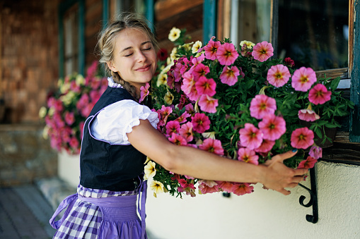 Teenage girl wearing Austrian and south German traditional dress - Dirndl.
The girl is hugging beautiful potted flowers on the windows in the the Austrian house.
Sunny summer day.
Canon R5