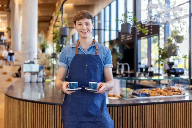 Photo of Smiling young barista serving two cups of coffee at office cafeteria
