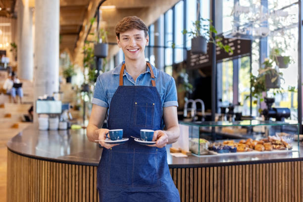 uśmiechnięty młody barista serwujący dwie filiżanki kawy w biurowej stołówce - cafeteria food service business zdjęcia i obrazy z banku zdjęć