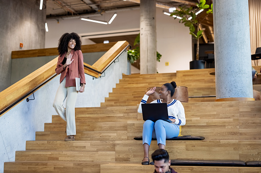 Woman walking downstairs and waving to a colleague sitting on steps at creative office. Female entrepreneur greeting coworker sitting on steps at office.