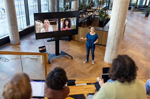Businesswoman discussing with team during video conference. Female talking with colleagues in office and two women connecting via video call seen on television set.