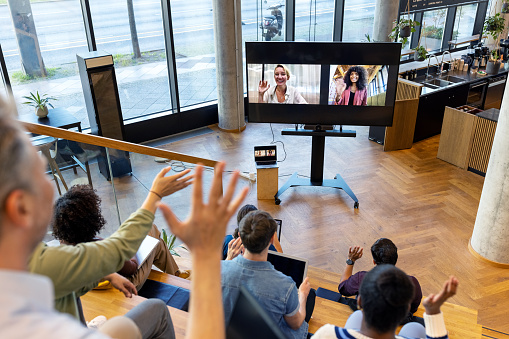 Startup business team having a hybrid conference meeting. Business people waving during video conference meeting with two female colleagues seen on large television screen.