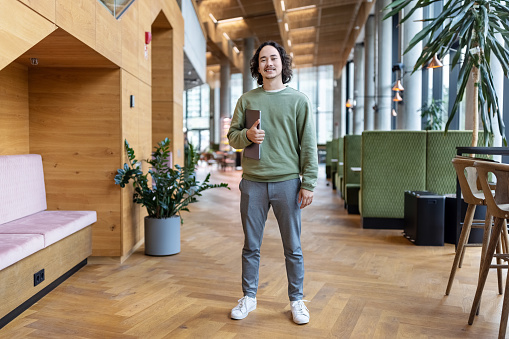 Full length portrait of confident young businessman standing in office lobby. Male entrepreneur holding laptop looking at camera at creative office.