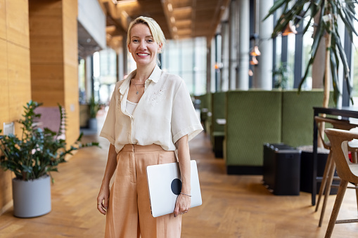 Portrait of a mature businesswoman standing in office lobby. Female entrepreneur with laptop looking at camera and smiling.