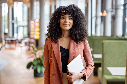 Portrait of confident African woman standing in office. Happy female entrepreneur with curly hair at office looking at camera.