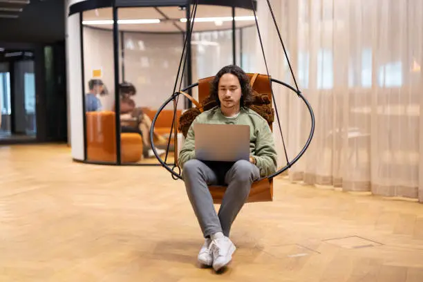 Young man working on laptop sitting on a swing chair in the coworking space. Young professional using laptop sitting in creative office break room.