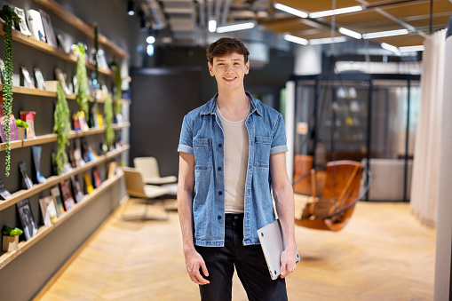 Portrait of a happy young man holding laptop standing at creative office break room. Smiling man in casuals standing in creative workspace.