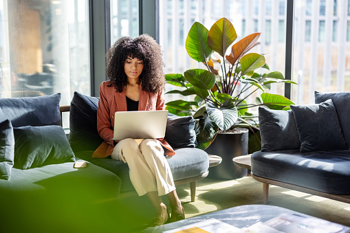 Young African woman working on laptop sitting at office lobby. Female entrepreneur with curly hair at office using laptop.