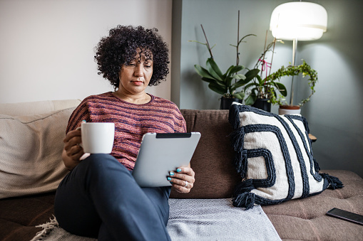 Multiracial mature woman holding digital tablet and coffee cup at home. She is surfing the net.