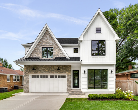 Oak Park, IL, USA - AUGUST 17, 2020: A new, white modern farmhouse with a dark shingled roof and black windows. The left side of the house is covered in a rock siding.