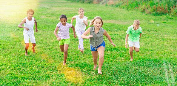 Portrait of international group of happy preteen boys and girls running on green lawn in city park on summer day