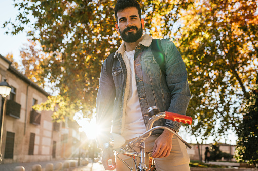 Portrait of a young man in denim jacket and backpack waiting on top of his vintage classic bicycle at sunset.