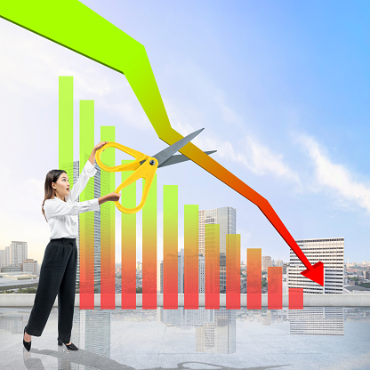 Asian businesswoman holding scissors to cut decrease arrow on the graph with cityscape background
