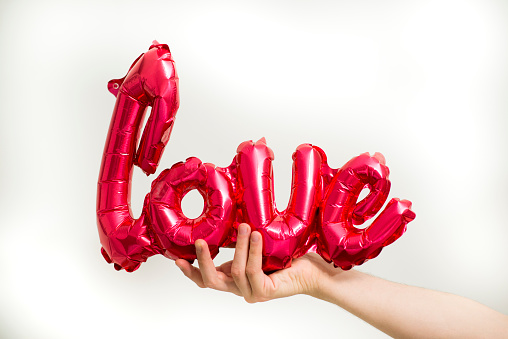 Anonymous young man's hand holding out a red, foil inflated balloon in the shape of the word love for Valentines day.