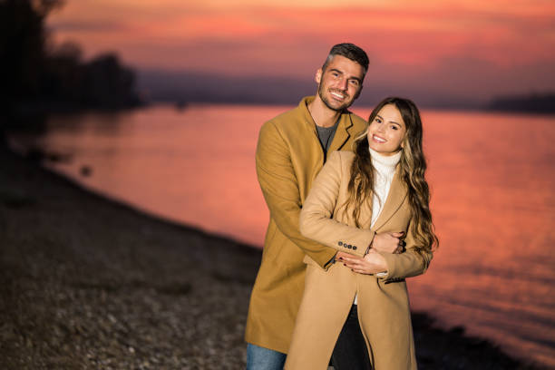 Beautiful young couple embracing while walking together next to river shore during sunset on autumn day. They are looking at camera.