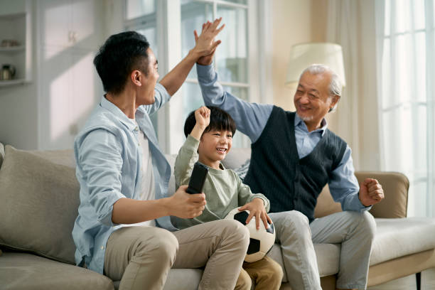 asian son father grandfather watching soccer game on TV together at home stock photo